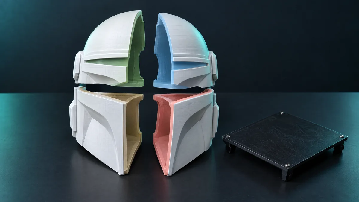 Capacete impresso em 3D cortado em quatro partes, cada corte de uma cor, ao lado de uma mesa de impressora
